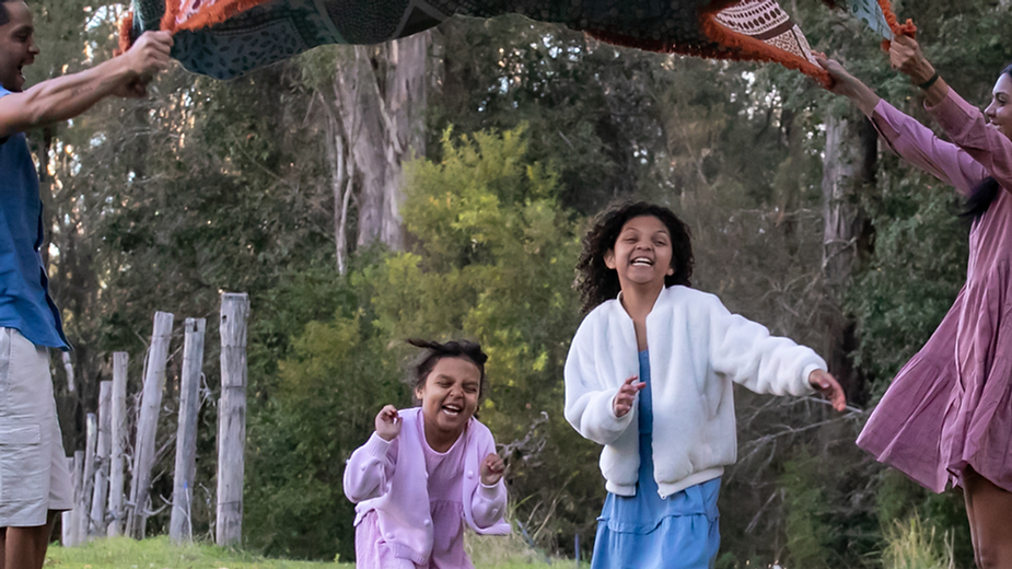 Two adults hold a patterned blanket above two laughing children in colorful dresses.