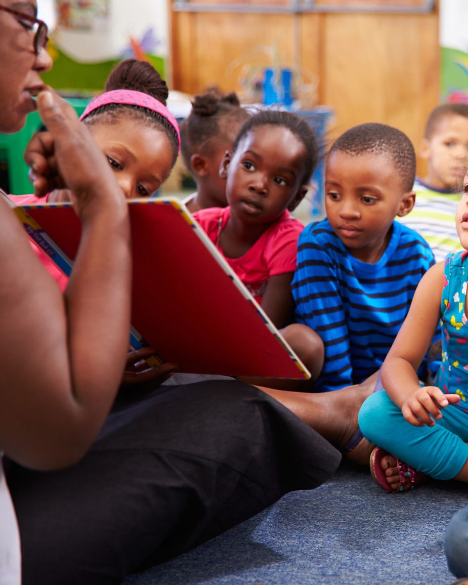 A group of children sit on a carpet, attentively listening to a person reading a book (speech)