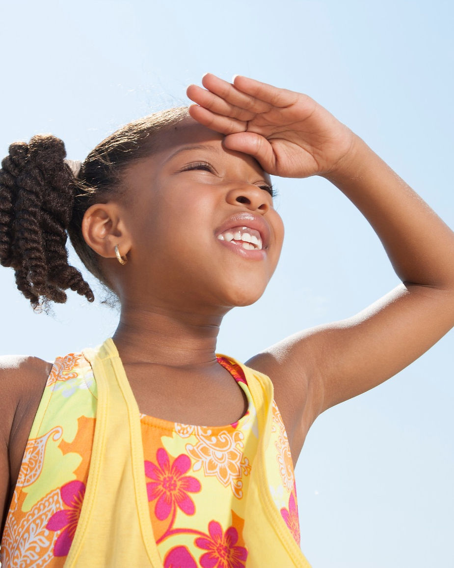 Girl in a colorful floral dress smiles, shielding her eyes with her hand under a bright blue sky.